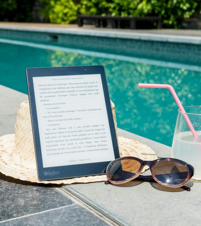 Sunny poolside scene with an e-reader, sunhat, sunglasses, and drink for ultimate relaxation.